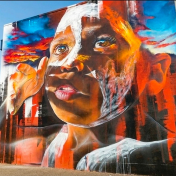 Large-scale street art mural of an Aboriginal person's face with traditional white face paint, painted in vivid oranges, blues and reds against a dramatic sky background